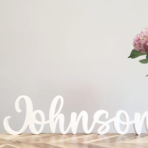 May include: White wooden sign spelling "Johnson" in a cursive font. The sign is placed on a wooden surface with a chevron pattern. A vase of pink hydrangeas is in the background.