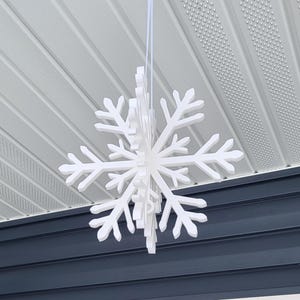May include: A white snowflake ornament hanging from a white ceiling. The snowflake is made of paper or foam and has a 3D design.