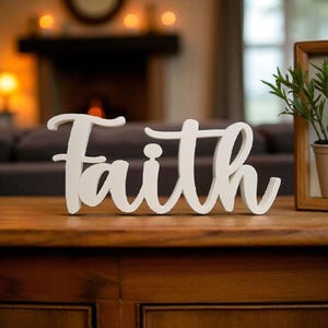 May include: A white wooden sign displaying the word "Faith" in a cursive font. The sign is placed on a wooden table, with a blurred background featuring a fireplace and framed plant.