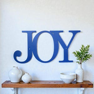 May include: A blue metal sign that spells out the word "JOY" is mounted on a white wall. Below the sign, a wooden shelf displays white vases, a bowl, and a vase with greenery. The sign is a decorative element.
