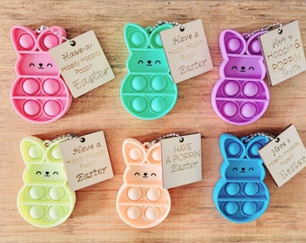 May include: Six colorful bunny-shaped pop-it fidget toys with attached keychains and wooden tags. The tags have different Easter-themed messages, including "Have a Hippity Hoppity Poppin Easter" and "Have a Hoppin & Poppin Easter".