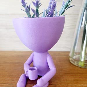 May include: A lavender plant sits in a purple planter shaped like a person holding a cup. The planter is made of a textured material and has a whimsical design.