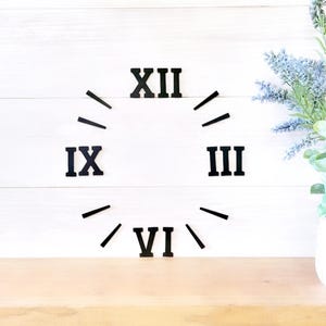 May include: Black Roman numeral clock face with the numbers XII, IX, III, and VI. The clock face is on a white wood background.
