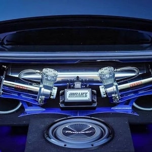 May include: The trunk of a blue car with a custom air ride system. The system includes two air compressors, an air tank, and a subwoofer. The air compressors are mounted on blue brackets and the air tank is labeled "Air Lift". The subwoofer is a Pioneer brand.