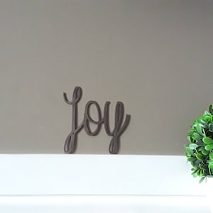 May include: A brown metal word art piece that spells out "joy" in a cursive font. The word is placed on a white surface against a neutral-colored wall. A green plant is on the right side of the image.