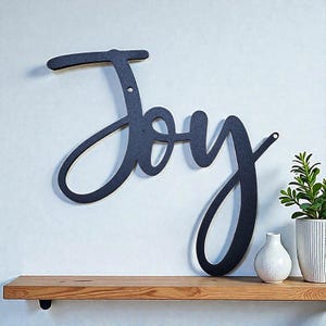 May include: A black metal wall art piece spelling out the word "Joy" in a cursive font. The word is mounted on a white wall above a wooden shelf. A small white vase and a potted plant are on the shelf.