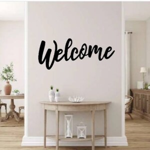 May include: A black metal welcome sign with a cursive font is mounted on a white wall above a light wood console table. The table has a shelf and is decorated with two white lanterns and a decorative piece.