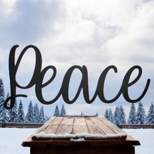 May include: A black metal sign with the word "Peace" in a cursive font, set against a snowy winter landscape with a wooden table in the foreground.