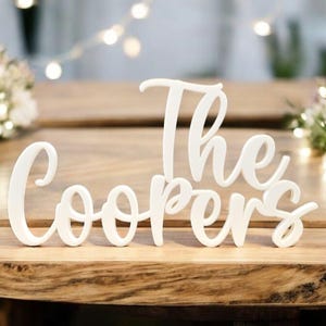 May include: White wooden sign that reads "The Coopers" in a cursive font. The sign is placed on a wooden table, with floral arrangements and string lights in the background. Perfect for wedding decor.