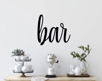 May include: A black cursive word "bar" on a white wall above a wooden countertop with a coffee maker, mugs, and a tiered tray with decorative items.