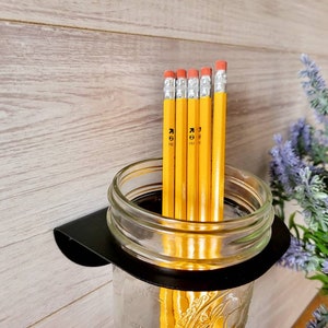 May include: A black metal wall mount with a clear glass jar holding multiple yellow pencils. The jar is attached to the wall mount with a black band.