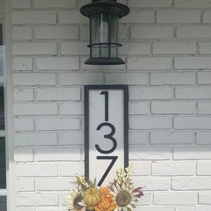 May include: Black and white house number sign with the number 137 on a white background. The sign is framed in black and has a black border. The sign is decorated with fall flowers and a small pumpkin.
