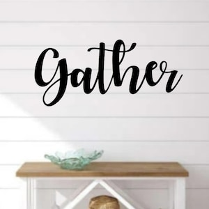 May include: Black cursive script sign with the word "gather" on a white wood wall above a wooden table with a glass bowl on top.