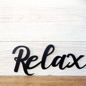 May include: Black cursive word "Relax" on a white and brown wooden background. The word is in a stylish, flowing font. The background has a rustic, farmhouse style.