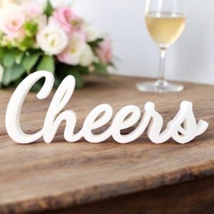 May include: White wooden sign with the word "Cheers" in a cursive font. The sign is sitting on a wooden table.