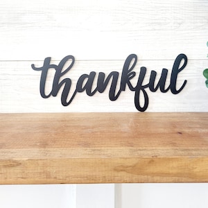 May include: Black metal sign with the word "thankful" written in a cursive font.