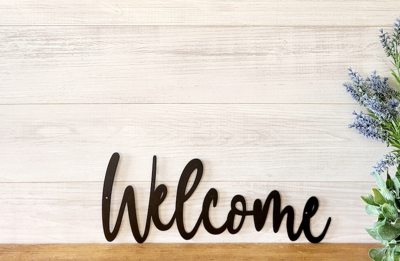 May include: Black metal welcome sign with a cursive font on a white wood background.