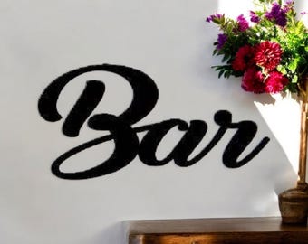 May include: Black cursive script sign that reads "Bar" on a white wall. A bouquet of pink and purple flowers is in a vase to the right of the sign.
