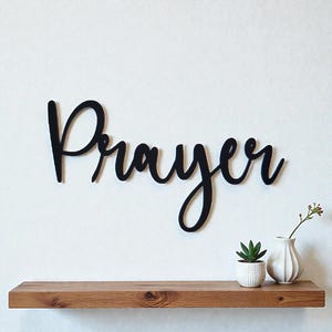 May include: Black cursive word art spelling "Prayer" mounted on a white wall. A wooden shelf below holds a small succulent in a white pot and a white vase with a sprig of greenery. The wall art adds a touch of faith-based decor.