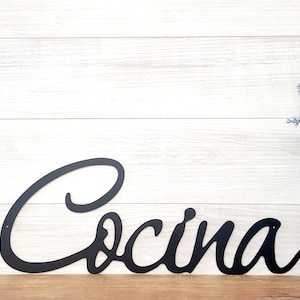 May include: Black metal sign with the word "Cocina" in a cursive font. The sign is on a white wood background.