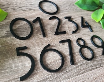 May include: Black numbers 0 through 9 in a modern, minimalist font style. The numbers are made of a thick, black material and are arranged in a random order on a wood surface.