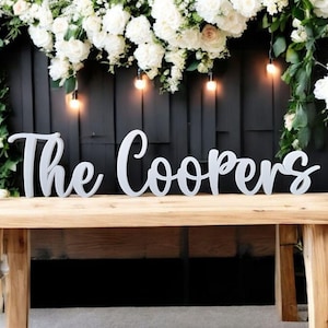 May include: White wooden letters spelling "The Coopers" on a wooden table with white flowers and string lights in the background.