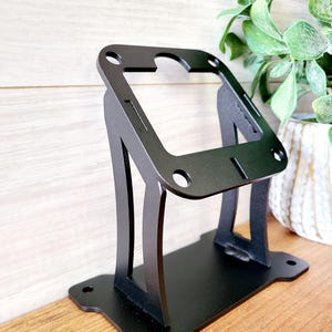 May include: Black metal wall mount stand with a rectangular opening. The stand has a decorative design with cutouts and a flat base.