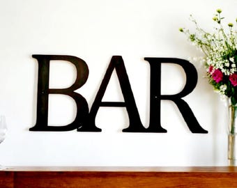 May include: Black metal letters spelling out the word "BAR" mounted on a white wall. A vase with white and pink flowers is on the right side of the image.