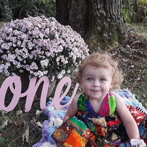 May include: A young child wearing a colorful patchwork dress sits in front of a pink wooden sign that says "one". The child is smiling and looking at the camera. The sign is surrounded by white flowers.
