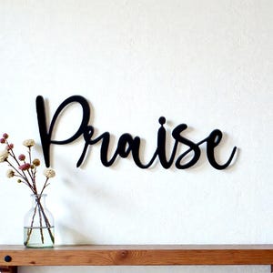 May include: Black wooden "Praise" sign in a cursive font, mounted on a white wall above a wooden shelf. A clear glass vase with flowers sits on the shelf. The sign is a decorative element.