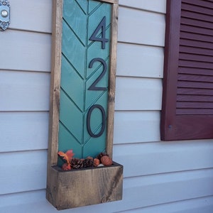 May include: A wooden house number sign with a green chevron pattern and black numbers reading "420". The sign has a small planter box underneath with fall-themed decorations.