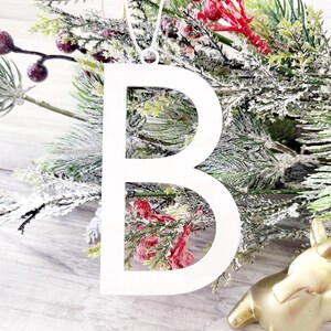May include: White metal ornament shaped like the letter B. The ornament is hanging from a white string and is surrounded by green and white branches with red berries.