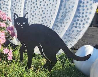 May include: Black metal silhouette of a cat standing in grass. The cat is facing the left side of the image. The cat is in front of a white pumpkin with blue polka dots.