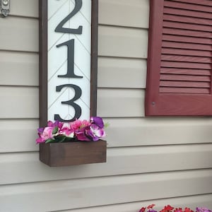 May include: A rectangular address sign with the numbers 213 in black against a white chevron pattern. The sign is framed in dark brown wood and has a small wooden planter box filled with pink and purple flowers. The sign is mounted on a beige siding wall.