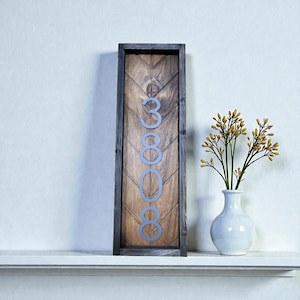 May include: A wooden address sign with the numbers 3808 in silver. The sign has a dark gray frame and a chevron pattern on the wooden background. A white vase with yellow flowers sits to the right.