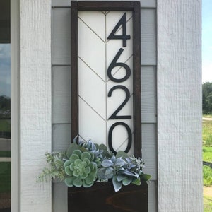 May include: A wooden sign with a white chevron pattern and black numbers 4620. The sign is mounted on a white wall with a brown planter box underneath filled with green succulents.