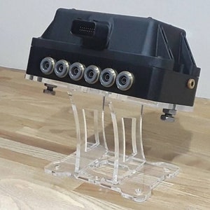 May include: A black electronic device with multiple ports and a clear acrylic stand. The device has a connector on the top and is mounted on the stand with screws.
