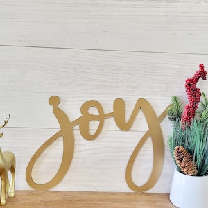 May include: A gold-colored, cursive "joy" sign is displayed against a white wooden background. Decorative gold deer figurines are on the left. A white vase holds greenery, red berries, and a pinecone on the right. Festive holiday decor.