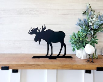 May include: Black metal moose silhouette wall decor with a rustic wooden shelf. The moose is standing on a metal bracket attached to the shelf.