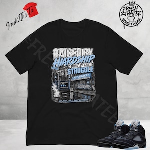 以下が含まれることがあります： RAISED BY HARDSHIP BUILT BY THE STRUGGLE、F2T MARKET、NOT BORN LUCKY, BUILT DIFFERENTというテキストと、地下鉄の入り口や店舗が描かれた都会の街並みのグラフィックプリントが施された黒の半袖Tシャツ。