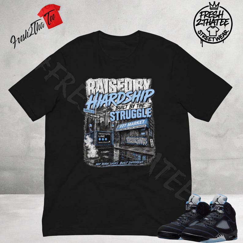 以下が含まれることがあります： RAISED BY HARDSHIP BUILT BY THE STRUGGLE、F2T MARKET、NOT BORN LUCKY, BUILT DIFFERENTというテキストと、地下鉄の入り口や店舗が描かれた都会の街並みのグラフィックプリントが施された黒の半袖Tシャツ。