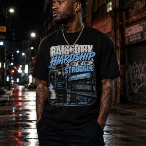 以下が含まれることがあります： RAISED BY HARDSHIP BUILT BY THE STRUGGLE、F2T MARKET、NOT BORN LUCKY, BUILT DIFFERENTというテキストがプリントされた黒の半袖Tシャツ。青と白のアクセントカラーで都会の街並みが描かれたグラフィックデザインが特徴です。