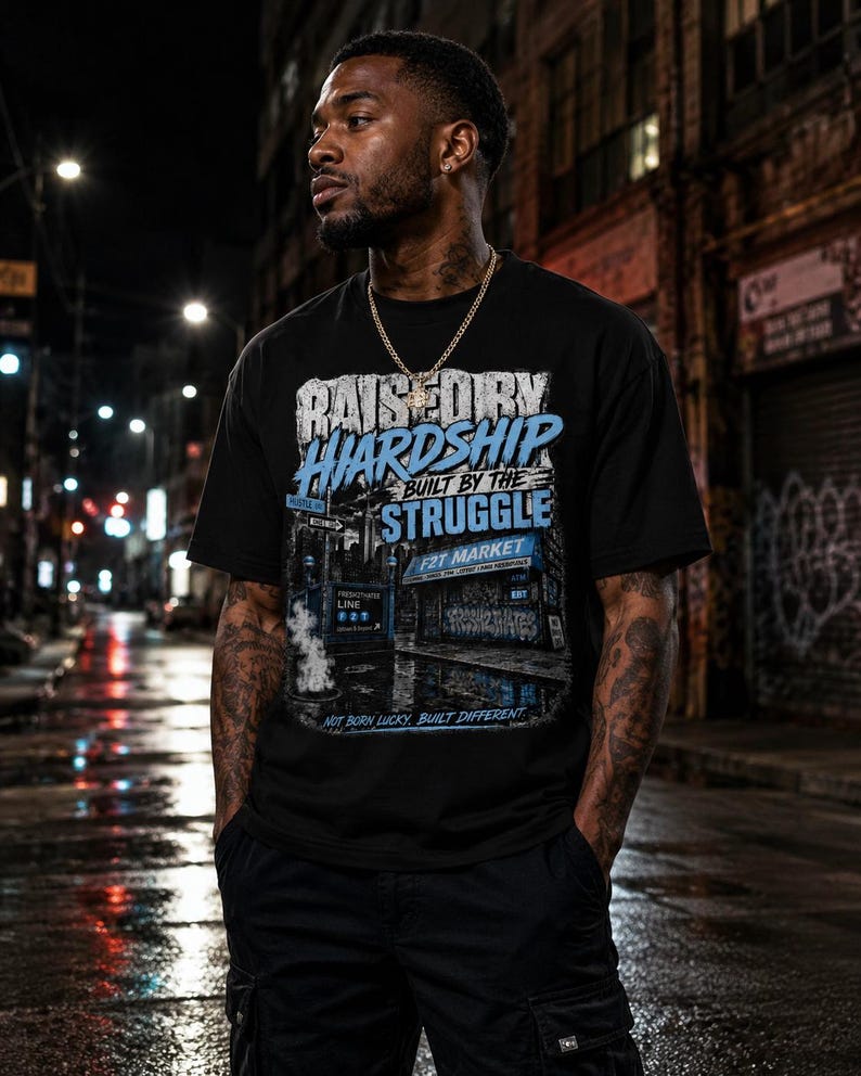 以下が含まれることがあります： RAISED BY HARDSHIP BUILT BY THE STRUGGLE、F2T MARKET、NOT BORN LUCKY, BUILT DIFFERENTというテキストがプリントされた黒の半袖Tシャツ。青と白のアクセントカラーで都会の街並みが描かれたグラフィックデザインが特徴です。