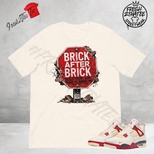 Pode incluir: Camiseta creme com um gráfico de um sinal de stop vermelho com as palavras "BRICK AFTER BRICK" e a frase "BUILT FROM WHAT TRIED TO BREAK ME". A camisa é combinada com tênis brancos e vermelhos.