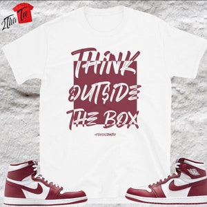 Puede incluir: Camiseta blanca con un gráfico granate que dice "Think Outside The Box" y un pequeño hashtag debajo. Dos zapatillas altas granate y blancas están a cada lado de la camiseta.