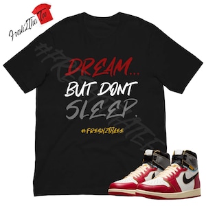 Pode incluir: Camiseta preta com o texto "Dream... But Don't Sleep." em letras vermelhas e cinzas. A camiseta também apresenta a hashtag "#Fresh2Thee" em amarelo. Um par de tênis vermelhos e brancos com um swoosh preto são mostrados no canto inferior direito da imagem.