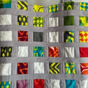 Op de afbeelding: Een draagtas met een rasterpatroon van veelkleurige stoffen vierkantjes in tinten geel, groen, blauw, rood, oranje en wit. De vierkantjes worden gescheiden door horizontale en verticale stroken van effen lichtgrijze stof met zichtbare stiksels.