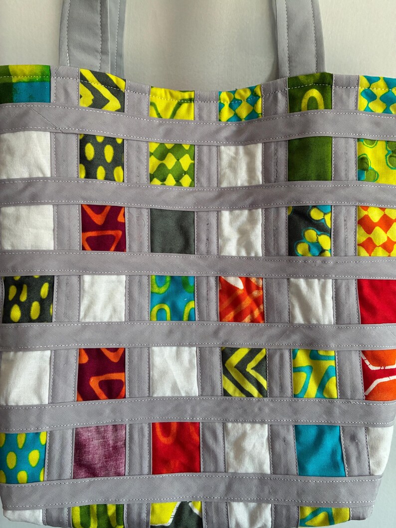Op de afbeelding: Een draagtas met een rasterpatroon van veelkleurige stoffen vierkantjes in tinten geel, groen, blauw, rood, oranje en wit. De vierkantjes worden gescheiden door horizontale en verticale stroken van effen lichtgrijze stof met zichtbare stiksels.