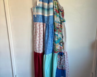 May include: A patchwork maxi dress with a variety of patterns and colors, including blue stripes, white hearts, and floral prints. The dress has a halter neckline with white ties and a ruffled lace hemline. The dress is hanging on a white hanger.
