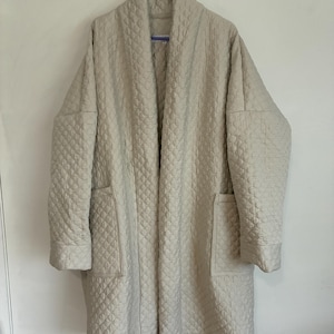 May include: A beige quilted long-sleeve coat featuring a shawl collar, an open front design, and two large patch pockets on the lower front. The garment is constructed with a repeating diamond-shaped stitched pattern throughout the fabric.